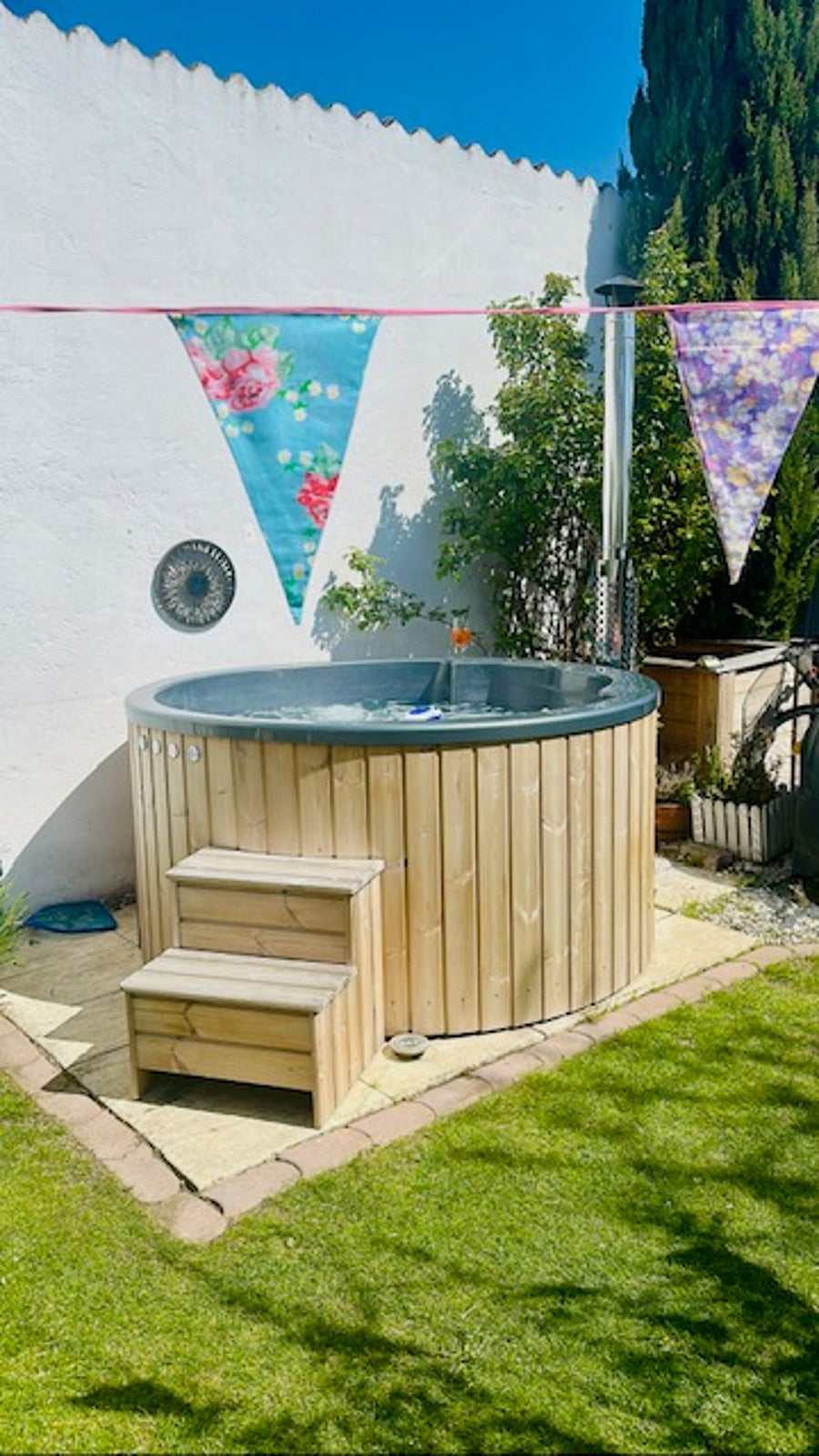 Runder Hot Tub mit Holzstiege und Wimpelgirlande im sonnigen Garten.